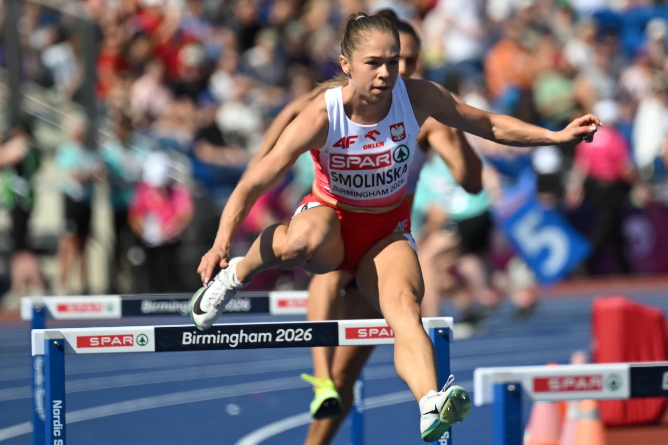 Izabela Smolińska, Birmingham 2026 Zawodniczka w biało-czerwonym stroju lekkoatletycznym przeskakująca przez płotek podczas biegu przez płotki na stadionie lekkoatletycznym, w otoczeniu innych rywalizujących biegaczek.