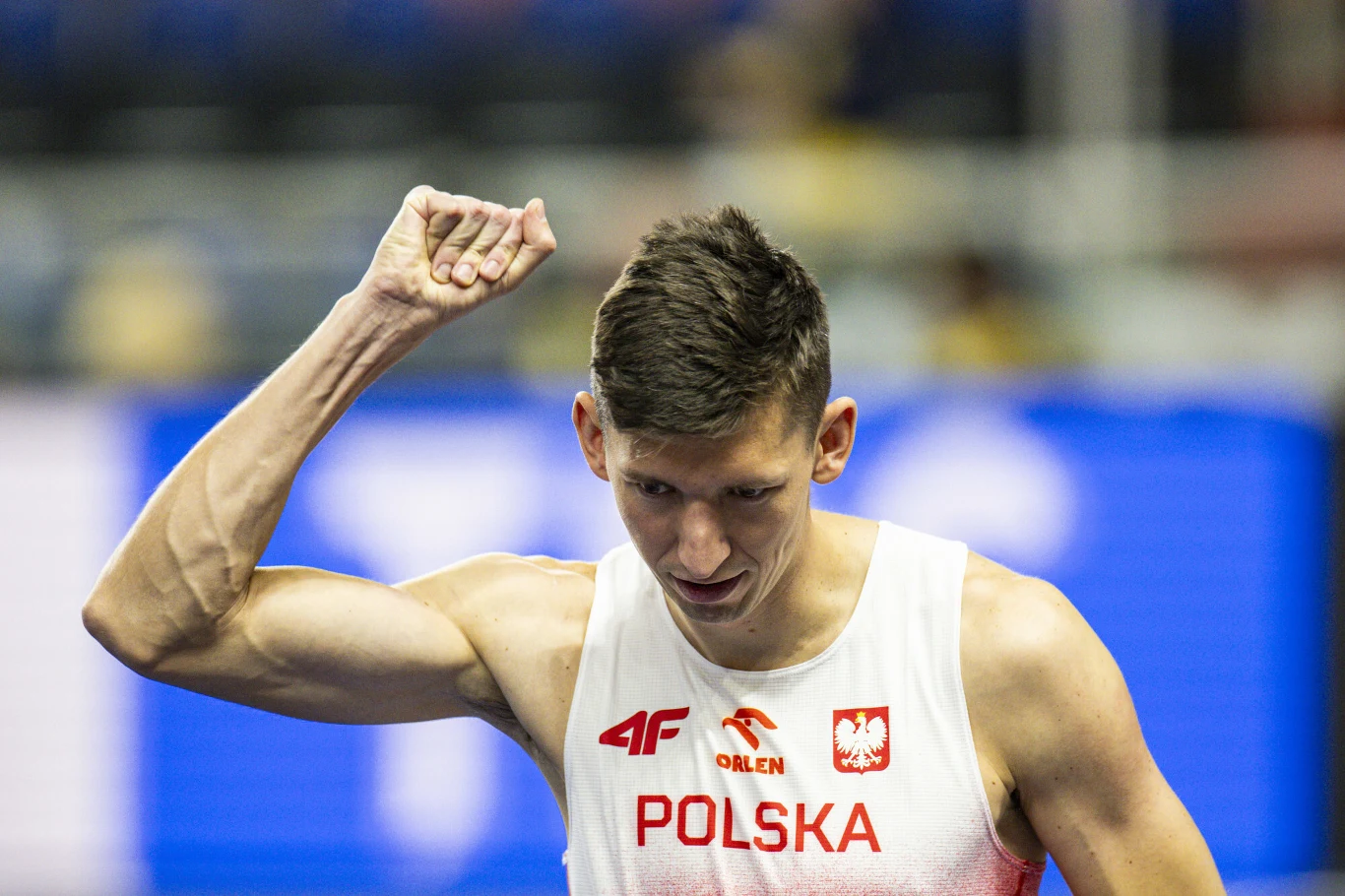 Lekkoatleta w białym stroju z napisem Polska i orłem na piersi unosi zaciśniętą pięść w geście triumfu na tle rozmytej hali sportowej.