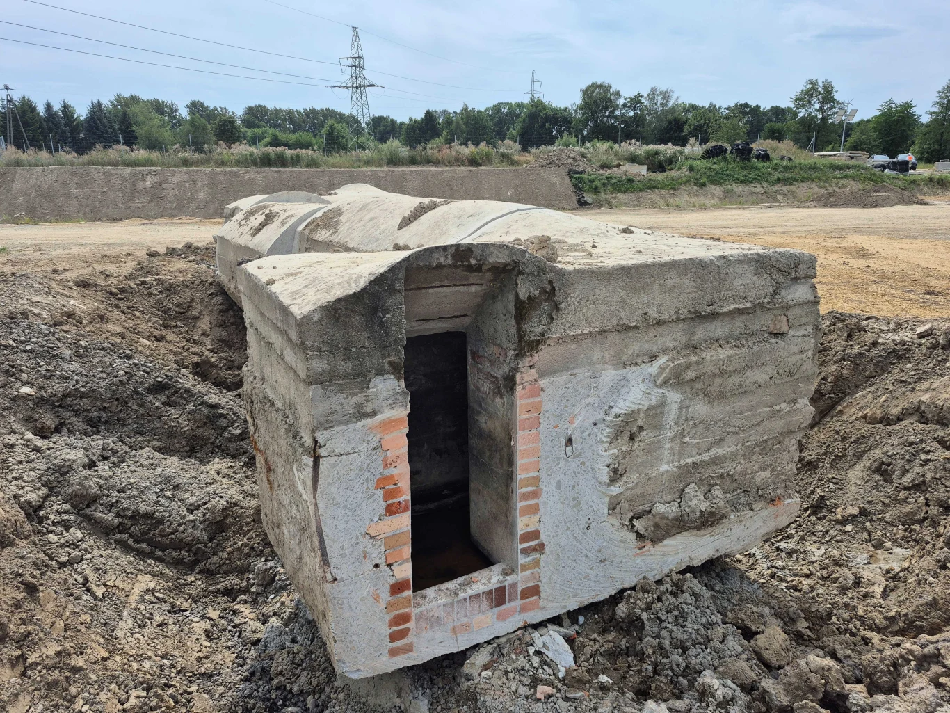 Betonowy bunkier z ceglaną obramówką wejścia częściowo wydobyty z ziemi, otoczony usypaną ziemią i gruzem, na tle terenów zielonych i słupów energetycznych.