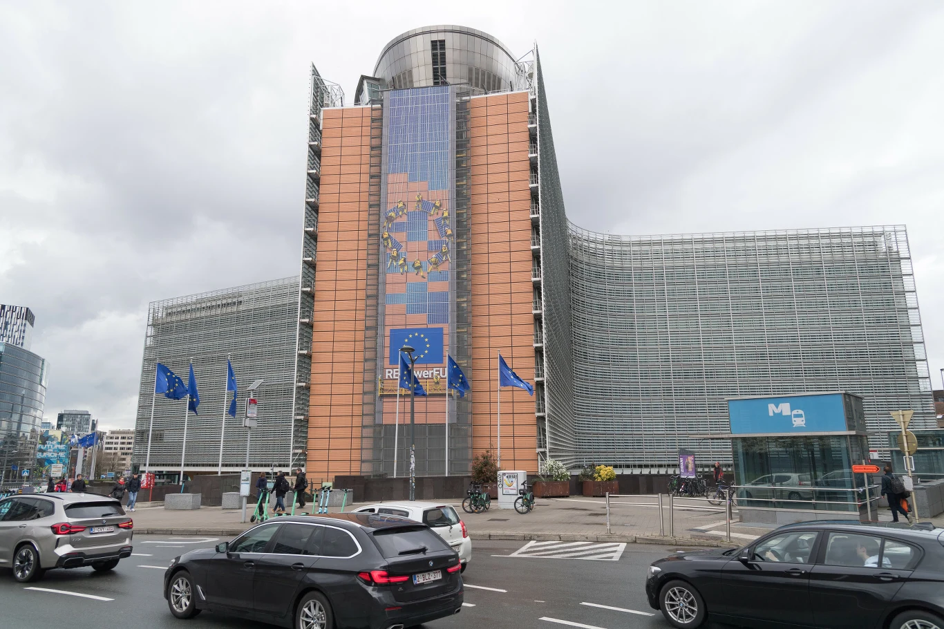 Siedziba Komisji Europejskiej przy Rondzie Schumana Nowoczesny budynek z charakterystyczną szklaną fasadą i centralnym pionowym bannerem z flagą Unii Europejskiej, otoczony flagami UE oraz samochodami poruszającymi się po ulicy przed obiektem.