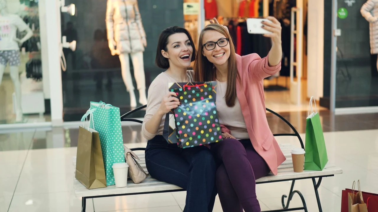 Dwie kobiety siedzą na ławce w galerii handlowej, trzymając kolorowe torby z zakupami i prezent oraz robiąc sobie wspólne selfie na tle witryn sklepowych.