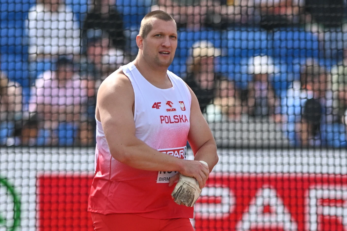 Wojciech Nowicki Lekkoatleta w czerwono-białym stroju z napisem Polska stoi na stadionie sportowym, trzymając kulę do pchnięcia kulą w dłoniach, w tle widoczne trybuny z widzami.