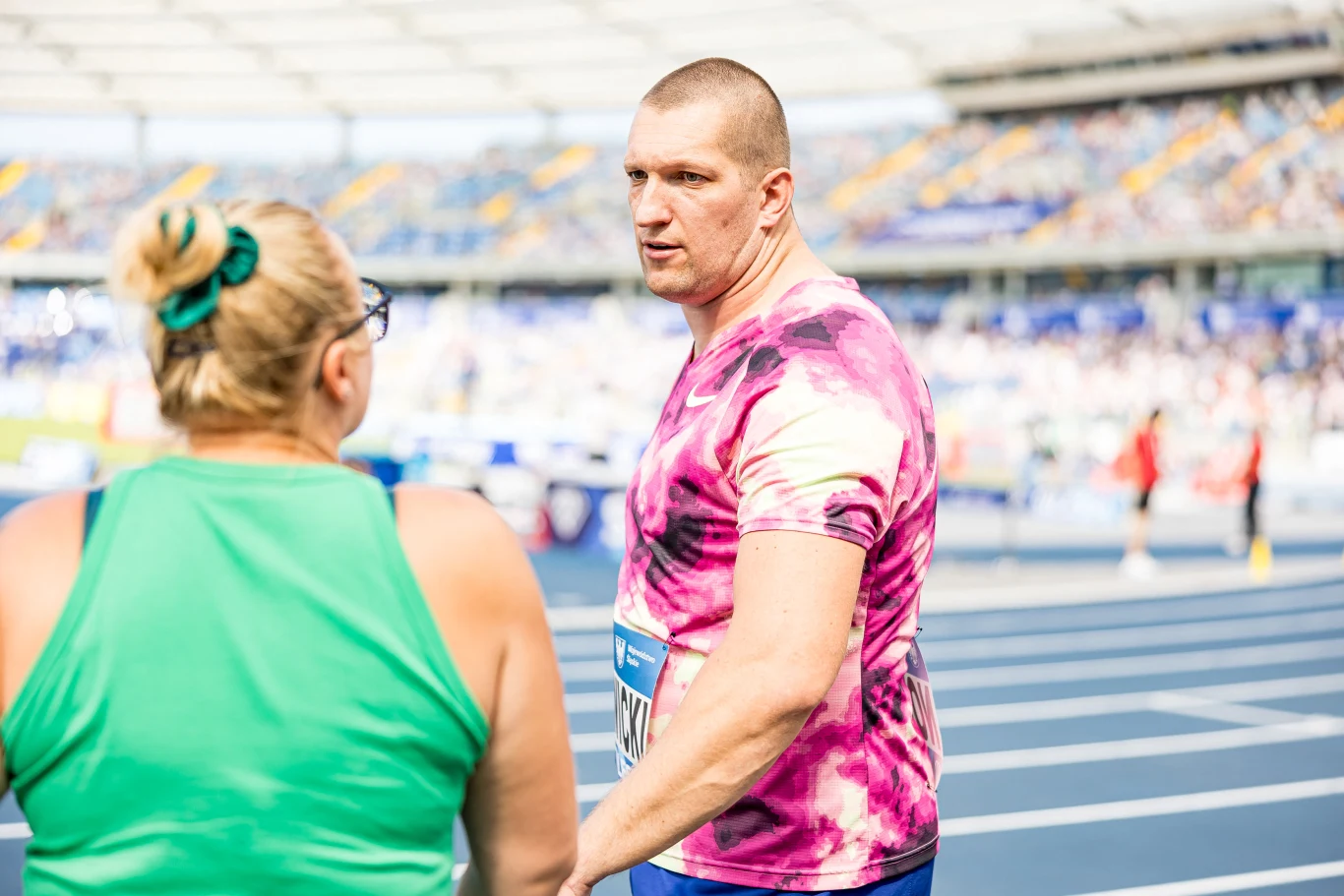 Wojciech Nowicki i trenerka Joanna Fiodorow Mężczyzna w różowej koszulce rozmawia z kobietą w zielonym stroju na stadionie lekkoatletycznym, w tle widoczne trybuny z kibicami.