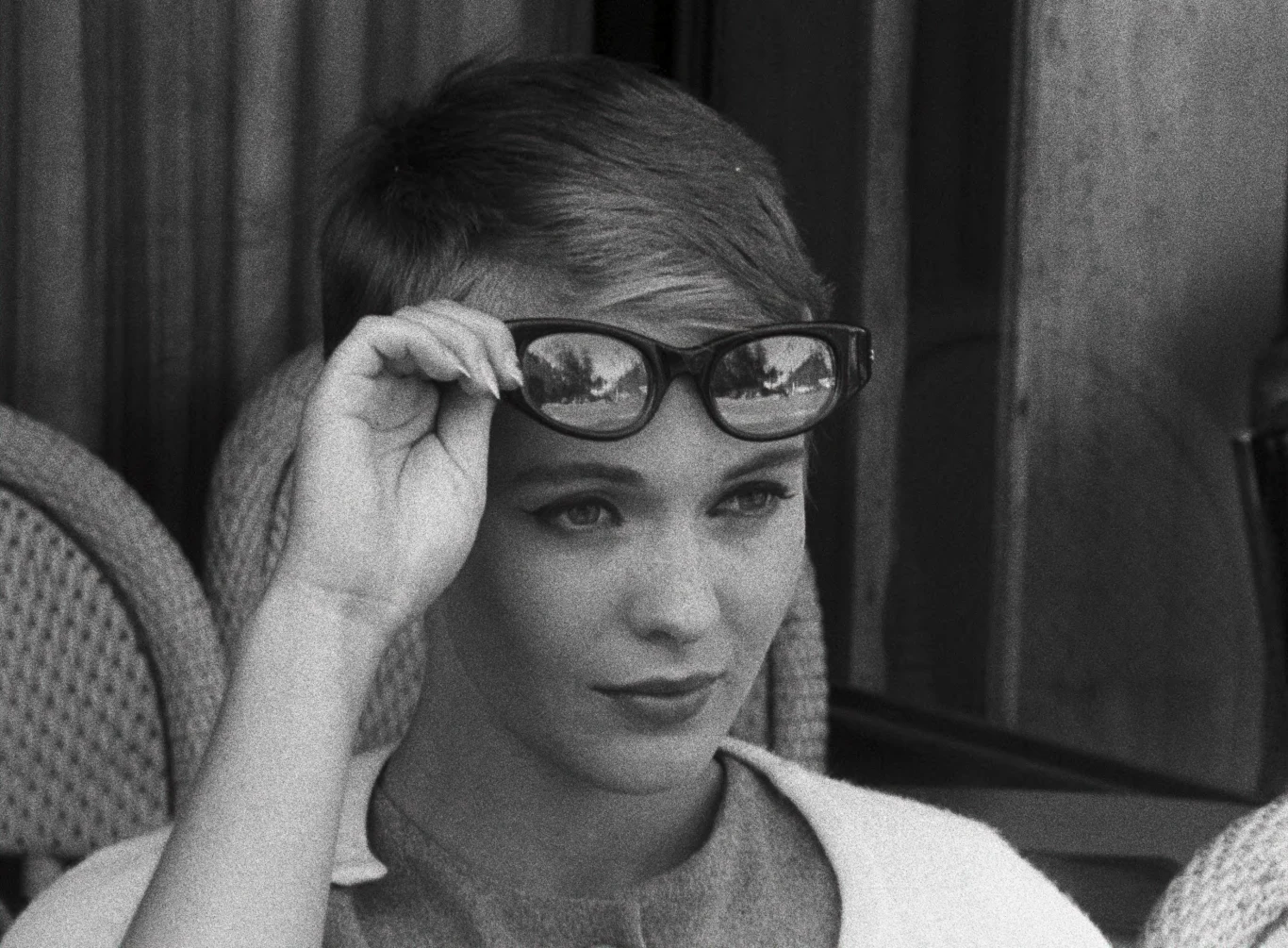 Jean Seberg w filmie "Do utraty tchu" (1960) / Past Perfect Młoda kobieta z krótkimi włosami, unosząca okulary przeciwsłoneczne do góry, spogląda uważnie przed siebie; za nią widoczne plecione krzesła ogrodowe.