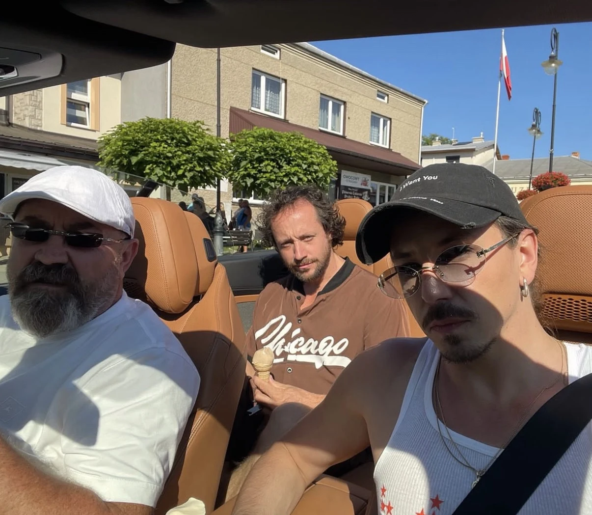 Michał Szpak z tatą i bratem Trzech mężczyzn siedzących w kabriolecie, jeden z nich trzyma loda, wszyscy patrzą w stronę aparatu; w tle budynki i polska flaga.