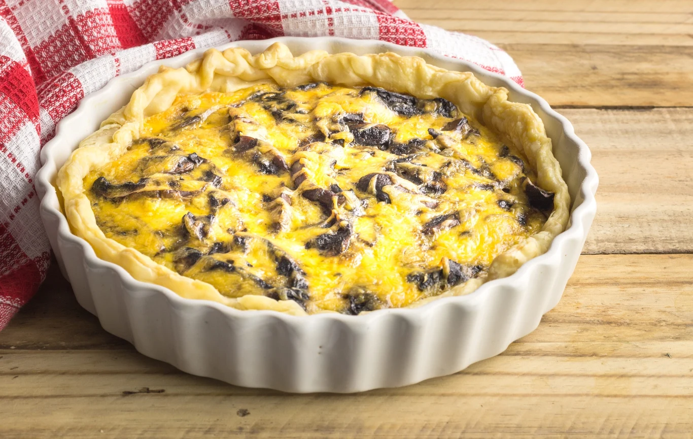 Quiche z pieczarkami i serem to rodzaj wytrawnej tarty z kruchego ciasta, masą jajeczno-śmietanową, grzybami i roztopionym serem Quiche z pieczarkami i serem w białej formie ceramicznej na drewnianym blacie, widoczne złociste brzegi.