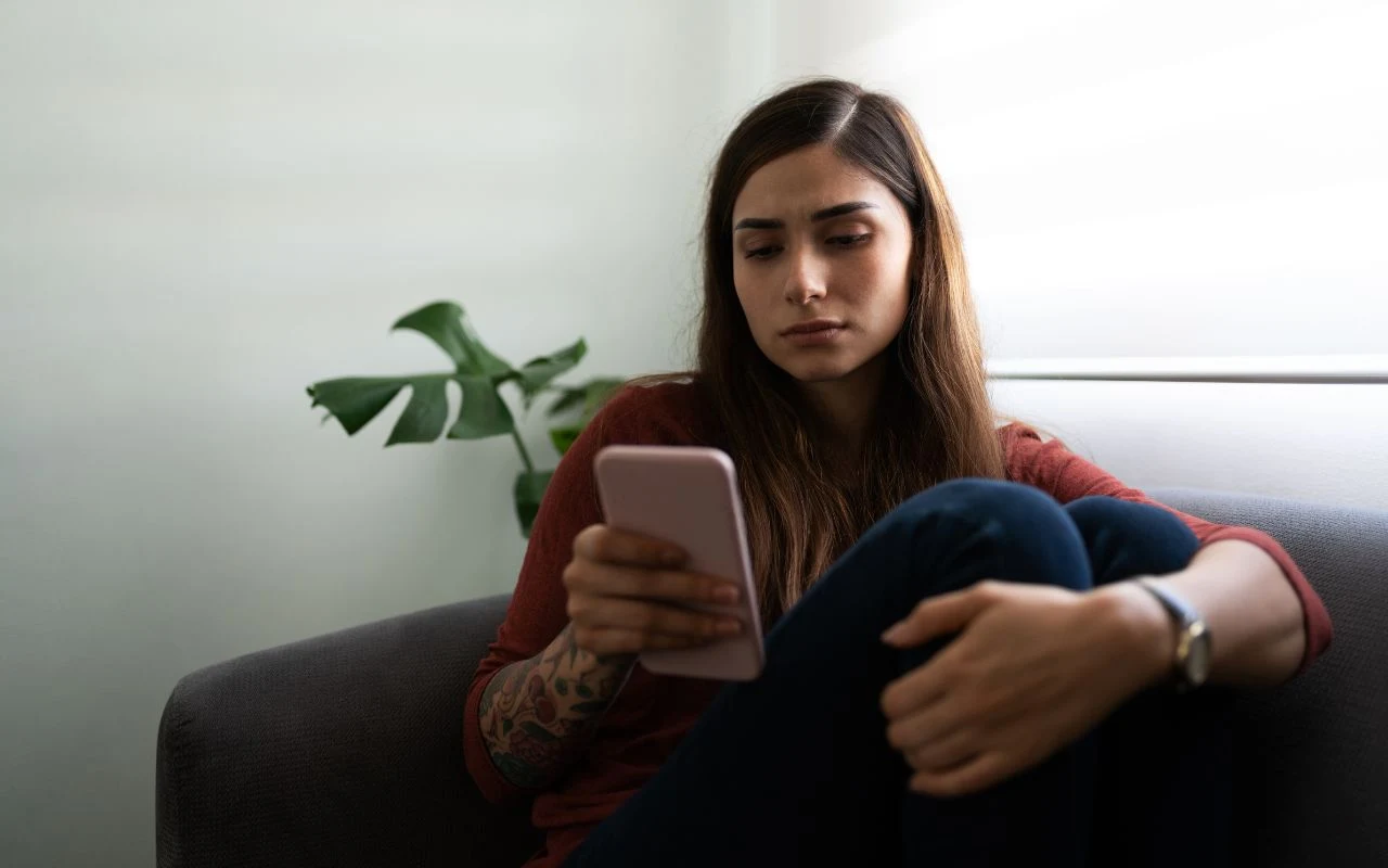 Młoda kobieta o długich ciemnych włosach siedzi na kanapie, trzymając w dłoni smartfon i patrząc na ekran z poważną miną.