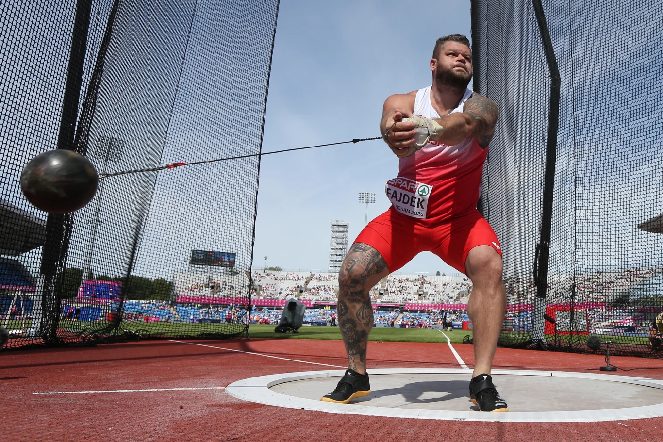 Paweł Fajdek Lekkoatleta w czerwono-białym stroju przygotowuje się do rzutu młotem na stadionie lekkoatletycznym, trzymając ciężki młot obiema rękami i koncentrując wzrok podczas dynamicznego ruchu w klatce rzutowej.