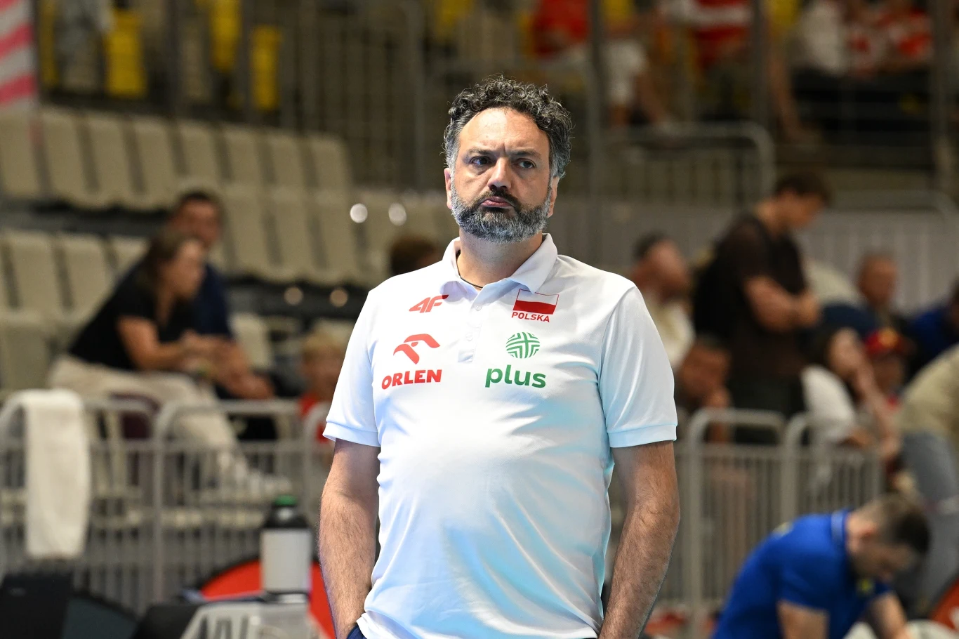 Stefano Lavarini, selekcjoner reprezentacji Polski siatkarek Trener siatkarski w białej koszulce reprezentacji Polski stoi przy boisku podczas meczu, skoncentrowany, z profesjonalnym wyrazem twarzy; w tle trybuny z kibicami.