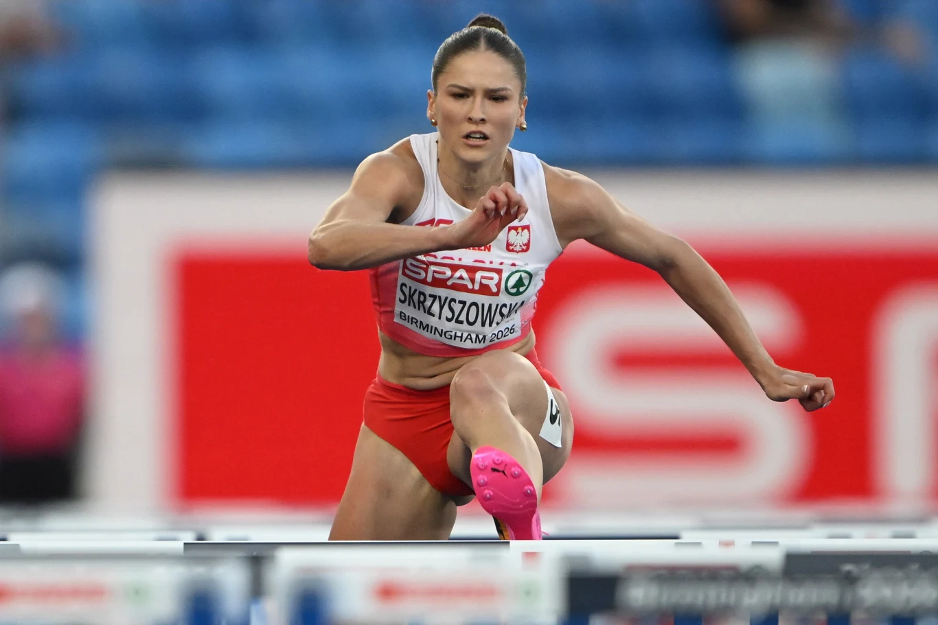 Pia Skrzyszowska, Birmingham 2026 Polska lekkoatletka w biało-czerwonym stroju przeskakuje przez płotek podczas biegu z przeszkodami na stadionie sportowym.