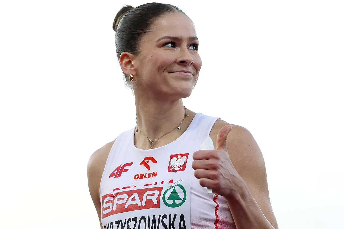 Pia Skrzyszowska, Birmingham 2026 Lekkoatletka w biało-czerwonym stroju z polskim godłem oraz logotypami sponsorów, unosi kciuk do góry i uśmiecha się, emanując pozytywną energią.