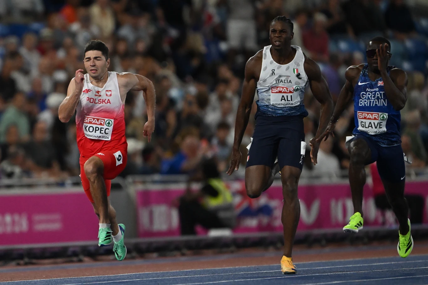 Trzech lekkoatletów w trakcie biegu na stadionie, każdy w stroju reprezentacyjnym swojego kraju; zawodnik z Polski po lewej, sportowiec z Włoch na środku i reprezentant Wielkiej Brytanii po prawej.