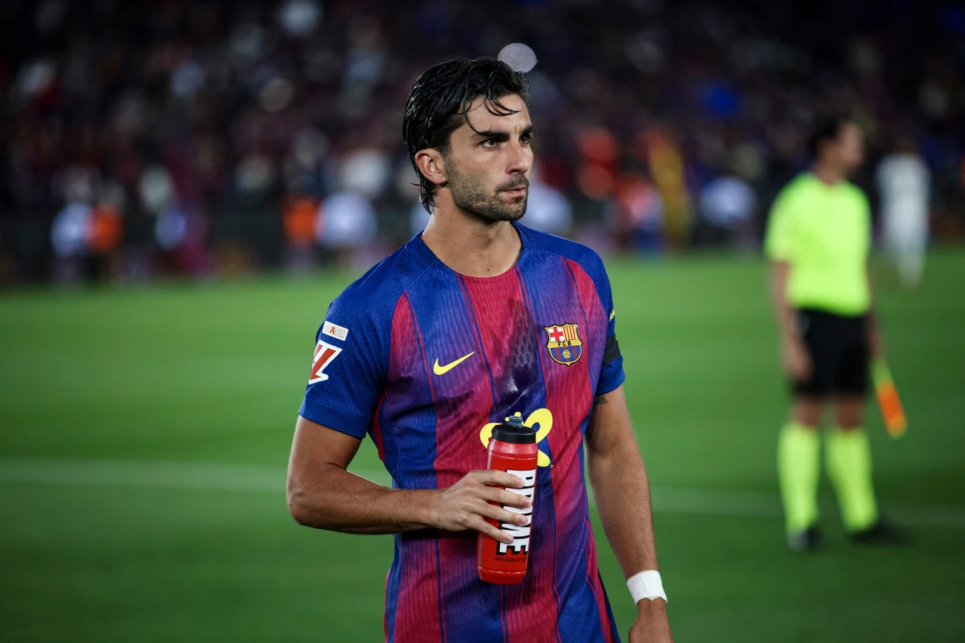 Ferran Torres Piłkarz FC Barcelony w granatowo-bordowej koszulce trzymający butelkę z wodą na murawie stadionu, w tle sędziowie w żółtych strojach.
