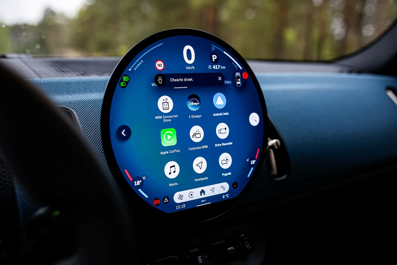 Okrągły panel multimedialny samochodu wyświetlający prędkość, wskaźnik paliwa, temperaturę oraz menu z ikonami aplikacji takich jak Apple CarPlay, Spotify i nawigacja.