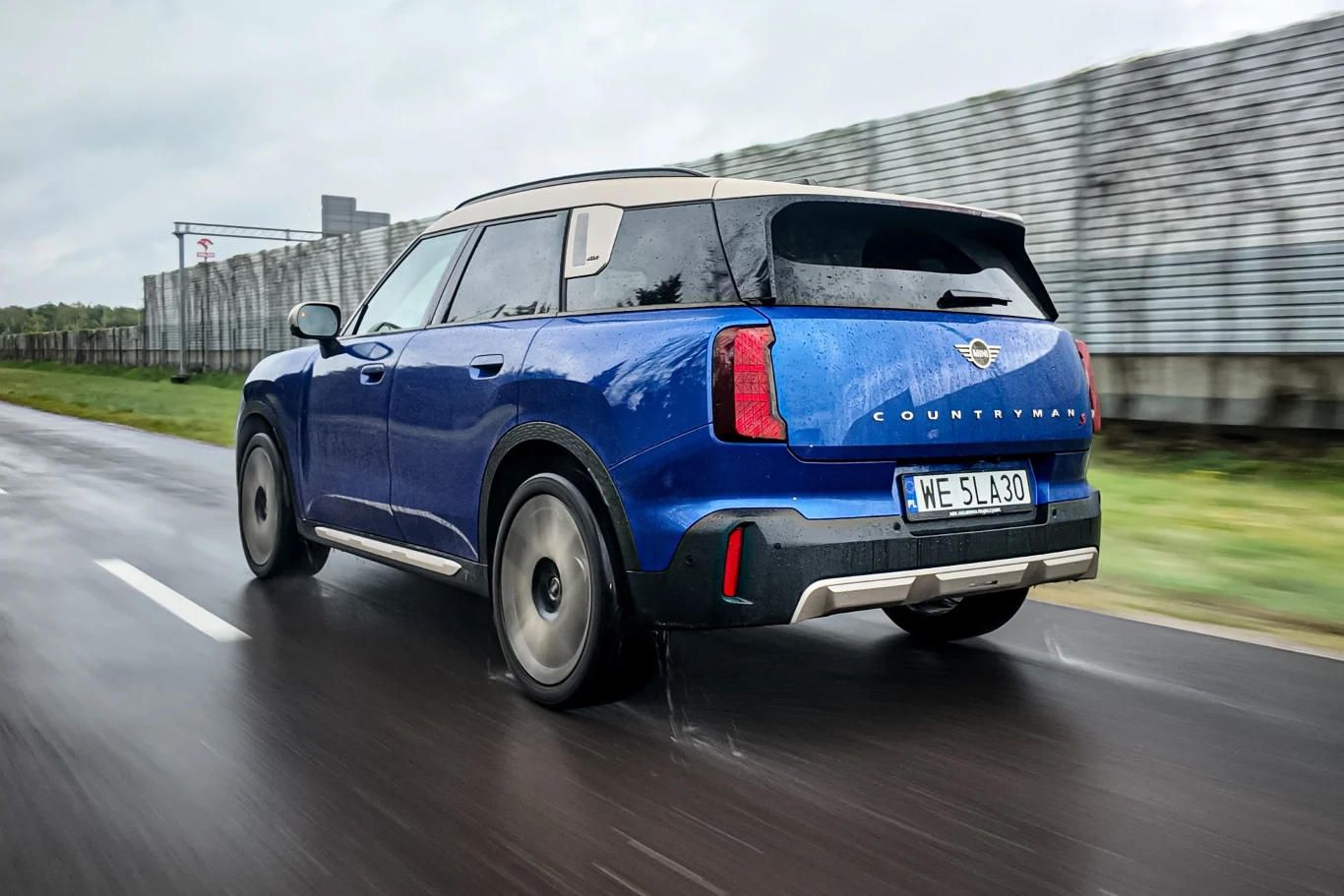 Niebieski SUV Mini Countryman jadący po mokrej nawierzchni drogą z wyraźnym oznakowaniem pasa oraz ogrodzeniem po prawej stronie.