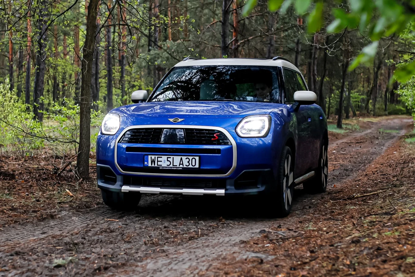 Niebieski Mini Countryman S z białym dachem jedzie leśną drogą otoczoną drzewami.