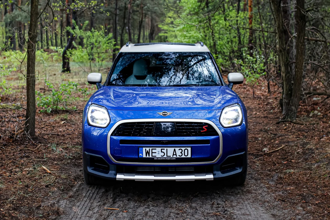 Niebieski Mini Countryman stoi na leśnej drodze otoczonej drzewami i zielonymi liśćmi, widok z przodu na maskę, reflektory i charakterystyczny grill samochodu.