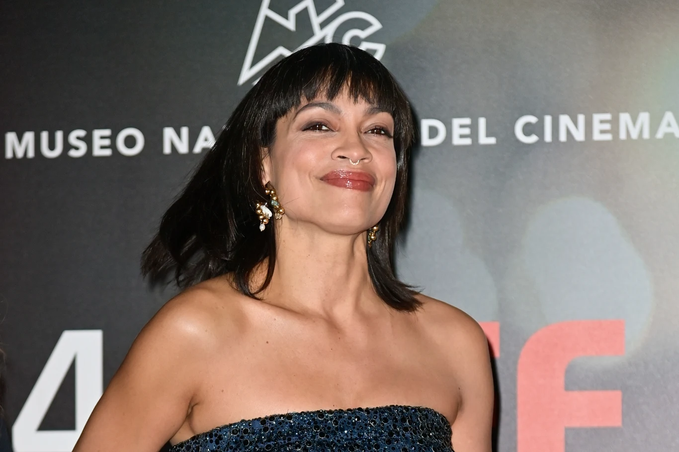 Rosario Dawson w 2024 roku Rosario Dawson w cekinowej, granatowej sukni bez ramiączek na tle czarnej ścianki z białymi napisami, 2024 rok.
