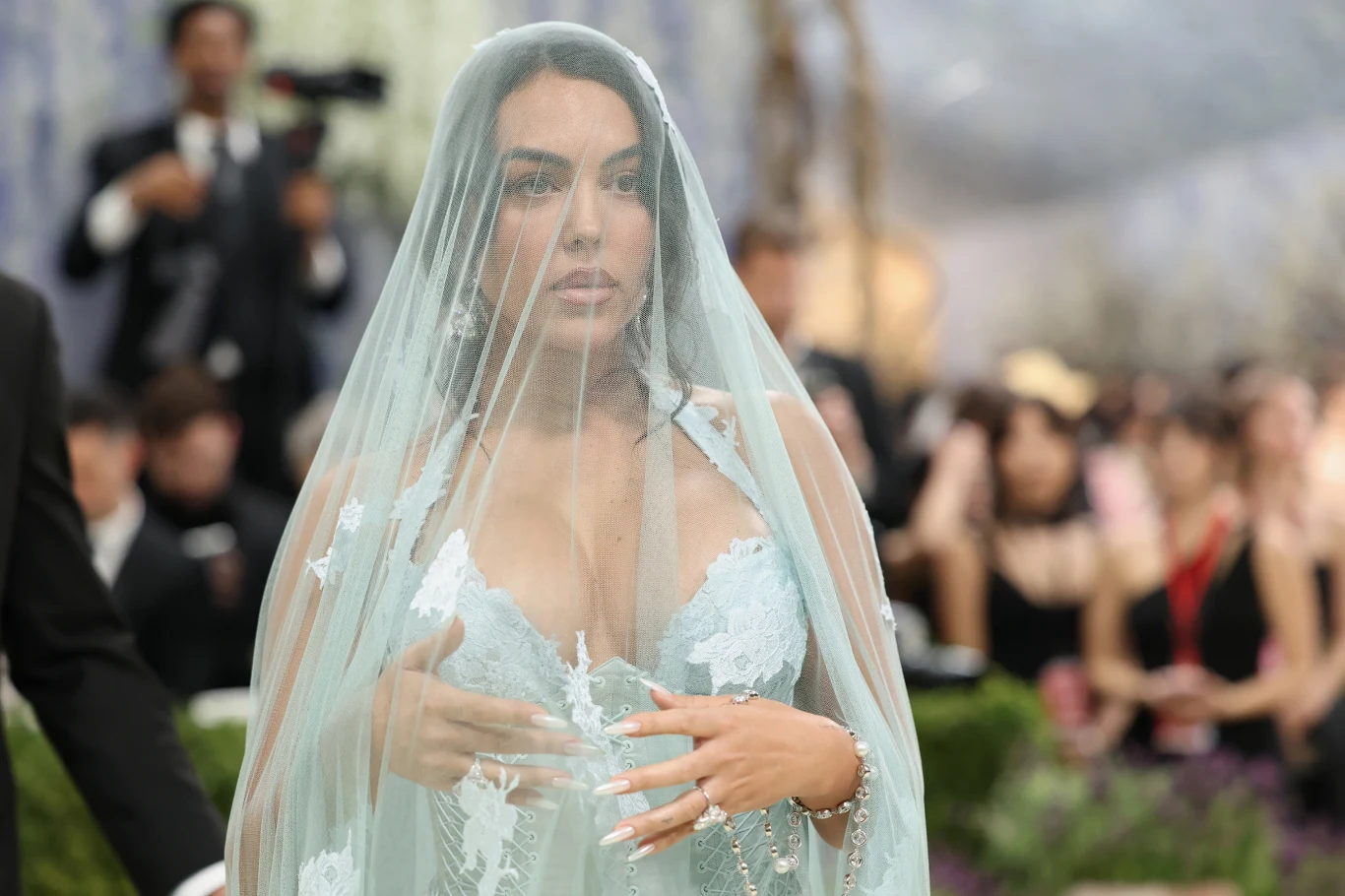 Kobieta w jasnoniebieskiej, koronkowej sukni z głębokim dekoltem, na głowie ma transparentny welon, w tle rozmyci uczestnicy wydarzenia w eleganckich strojach.