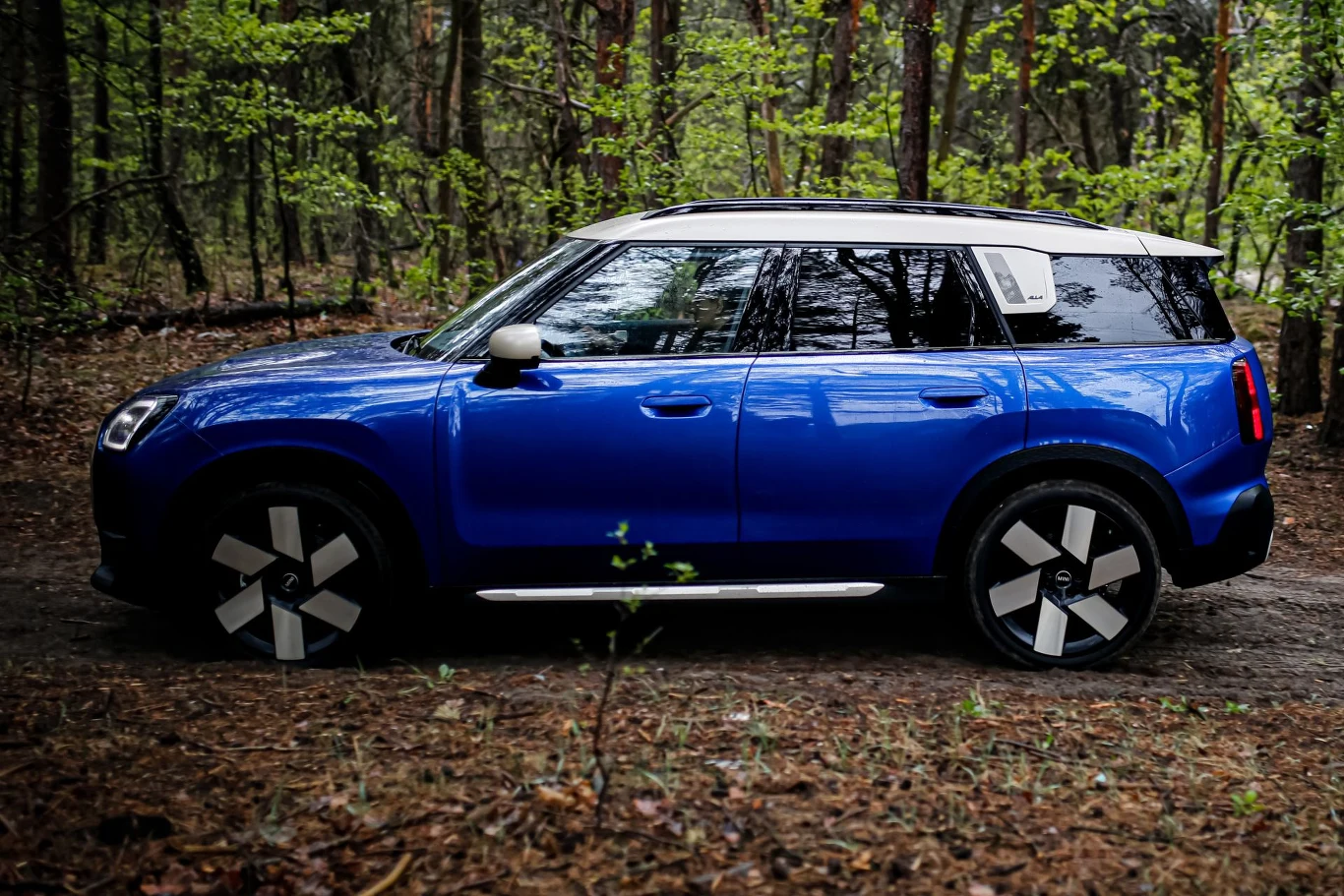 Niebieski SUV z kontrastującym białym dachem i charakterystycznymi felgami, zaparkowany na leśnej ścieżce wśród drzew.