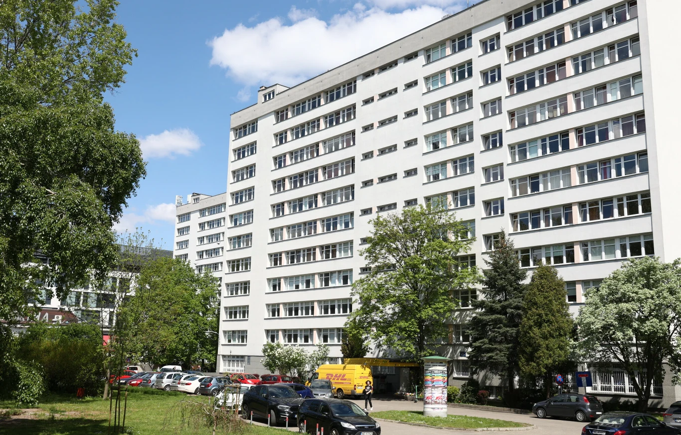 Wysoki blok mieszkalny z licznymi balkonami i dużymi oknami, przed którym znajduje się parking z zaparkowanymi samochodami oraz kilkoma drzewami i zielenią.