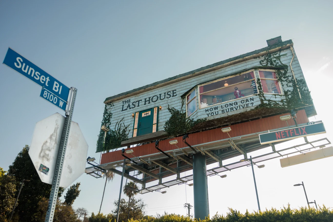 Trójwymiarowy billboard promujący produkcję Netflix, stylizowany na dom z roślinnością oplatającą elewację oraz dużym napisem „The Last House” i hasłem „How long can you survive?”; poniżej czytelny znak ulicy Sunset Blvd.