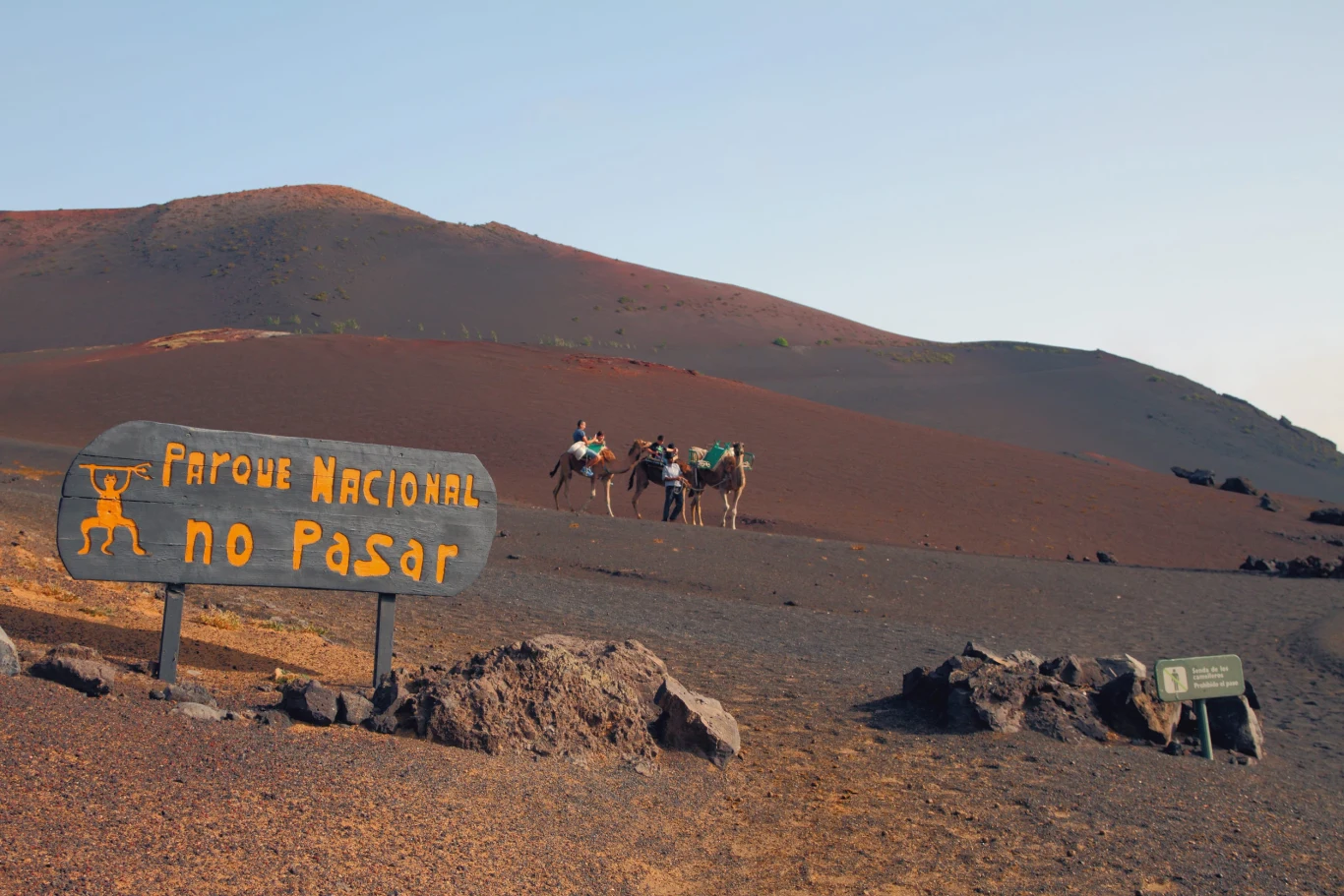 Timanfaya Trzy wielbłądy z jeźdźcami przemierzają skalistą, wulkaniczną pustynię, obok drewnianej tablicy z napisem zakazującym wstępu do Parku Narodowego.