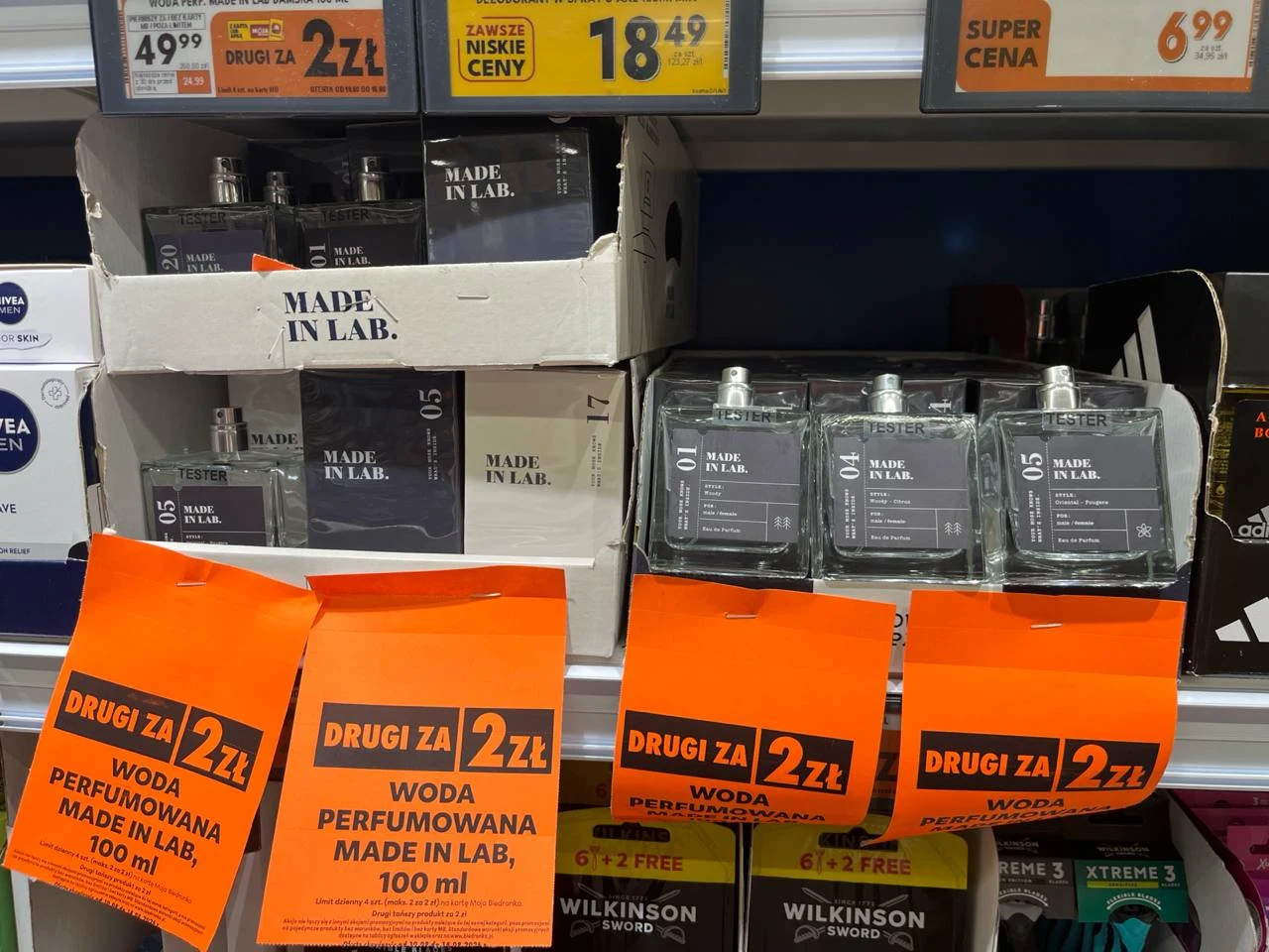 Drugie perfumy Made in lab za 2 zł. Sprawdziłam promocję w Biedronce Rzędy butelek wody perfumowanej marki Made in Lab ustawione na półkach, przed nimi wyraziste pomarańczowe etykiety promocyjne informujące o cenie drugiego produktu za 2 złote.