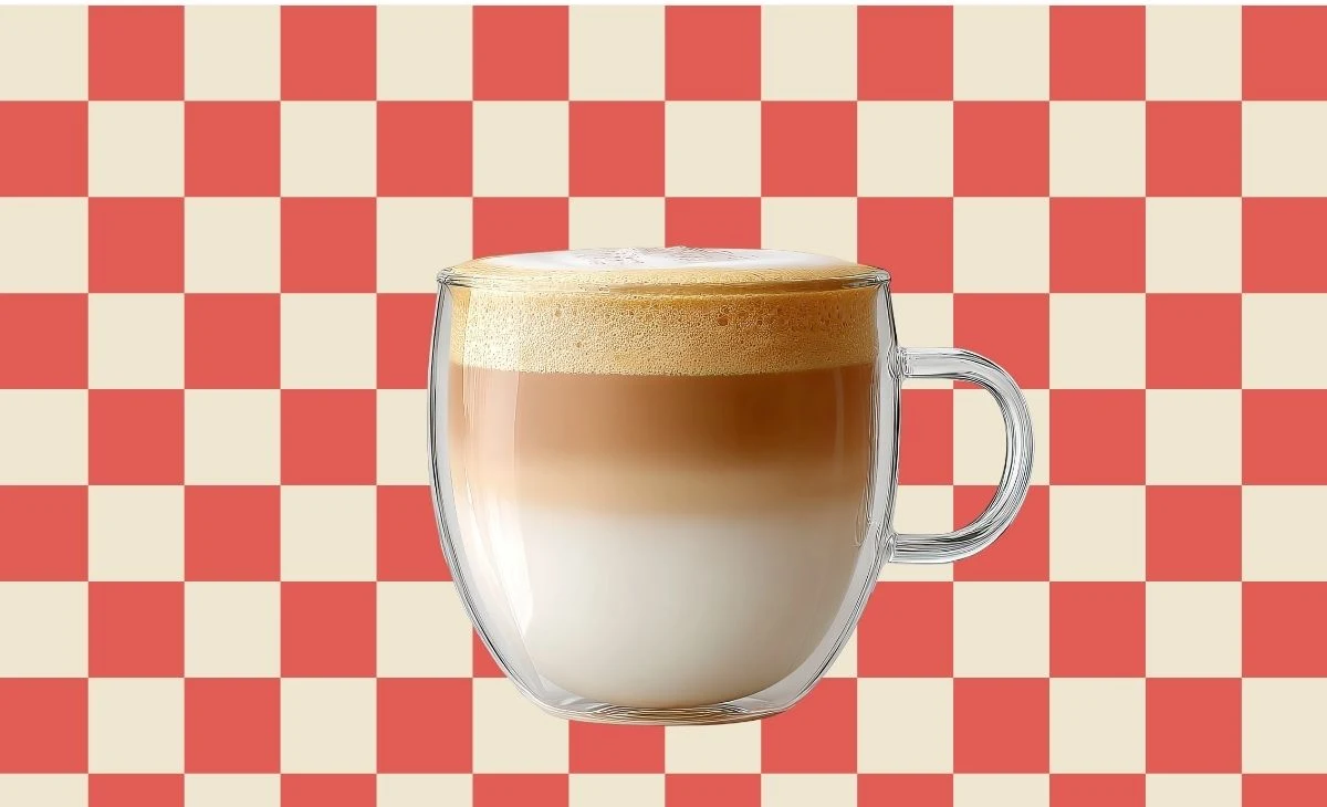 Zdecydowałaś się na latte? Lubisz, kiedy życie ma dobry smak Szklanka latte z wyraźnie oddzielonymi warstwami mleka, kawy i pianki ustawiona na tle czerwono-białej szachownicy.