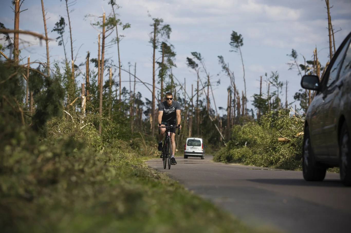 Rowerzysta jedzie pustą drogą otoczoną połamanymi i powalonymi drzewami, w tle widoczne dwa samochody, krajobraz nosi ślady zniszczeń po silnej burzy lub wichurze.