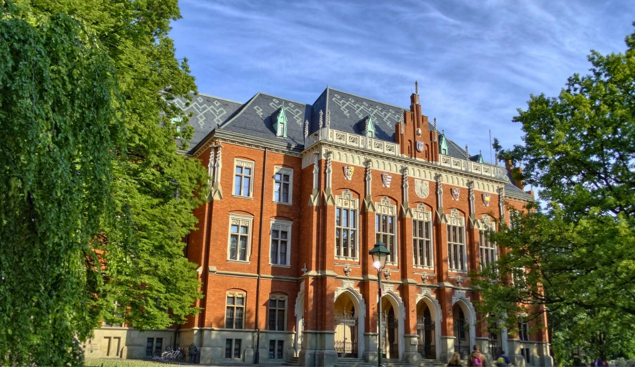 Wolne miejsca na studia 2026/2027 Ceglany budynek uczelni z ozdobnymi detalami i wysokimi oknami, otoczony drzewami pod błękitnym niebem