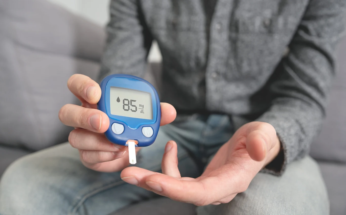 Osoba trzyma w dłoni glukometr z wynikiem 85 mg/dl, druga ręka gotowa do pobrania próbki krwi z palca.