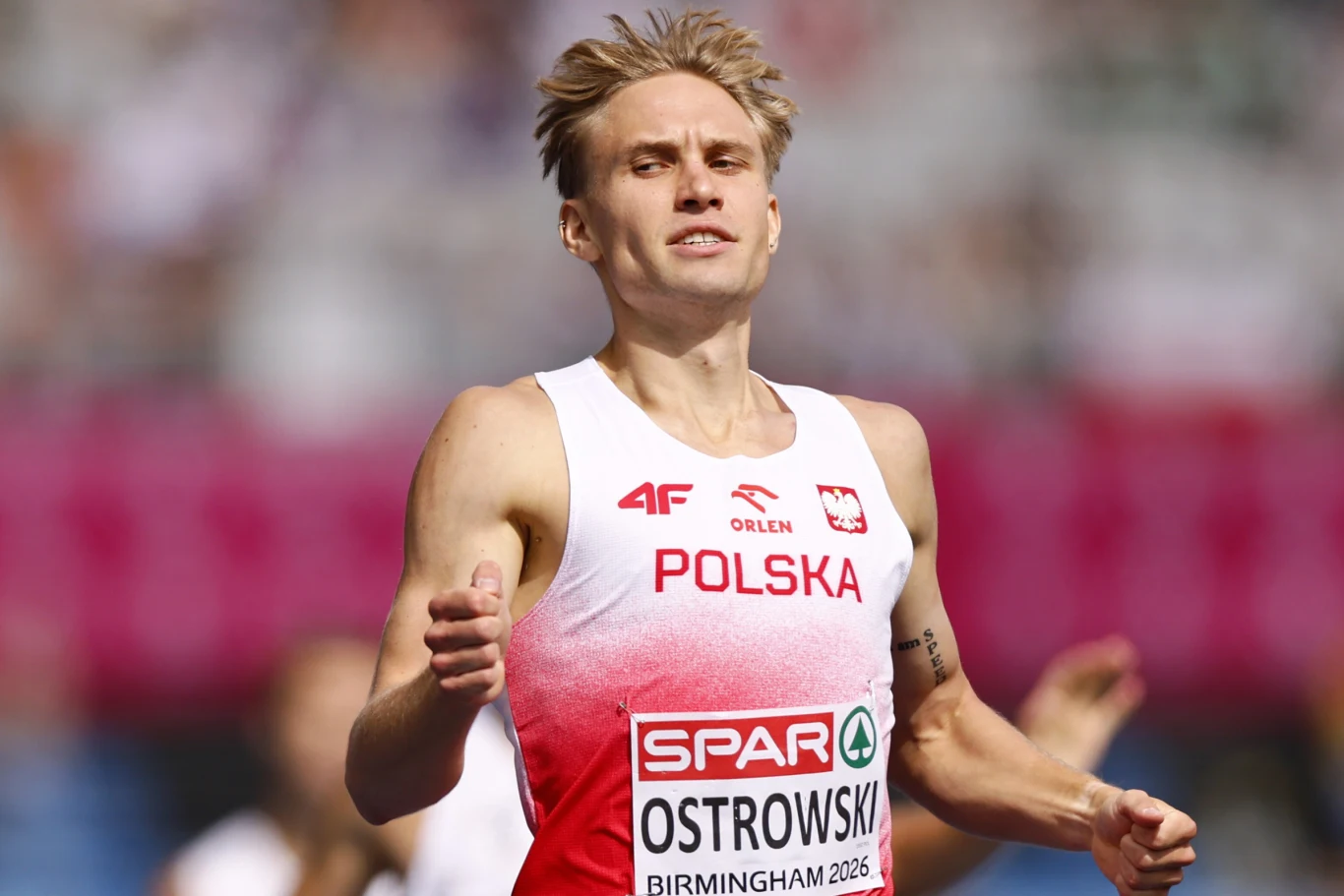 Polski lekkoatleta w stroju reprezentacyjnym podczas biegu na stadionie, skupiony w trakcie rywalizacji sportowej.