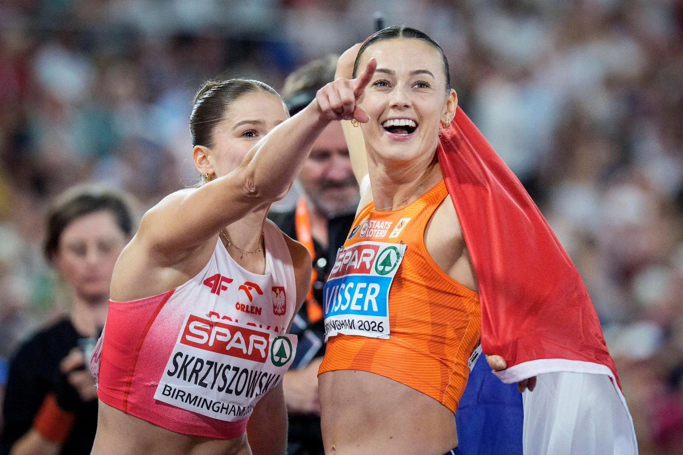 Pia Skrzyszowska i Nadine Visser Dwie lekkoatletki na bieżni, jedna z nich owinięta narodową flagą i uśmiechnięta wskazuje coś w oddali, obok niej druga sportsmenka również wyraziście reaguje na wydarzenia po ukończonym biegu.
