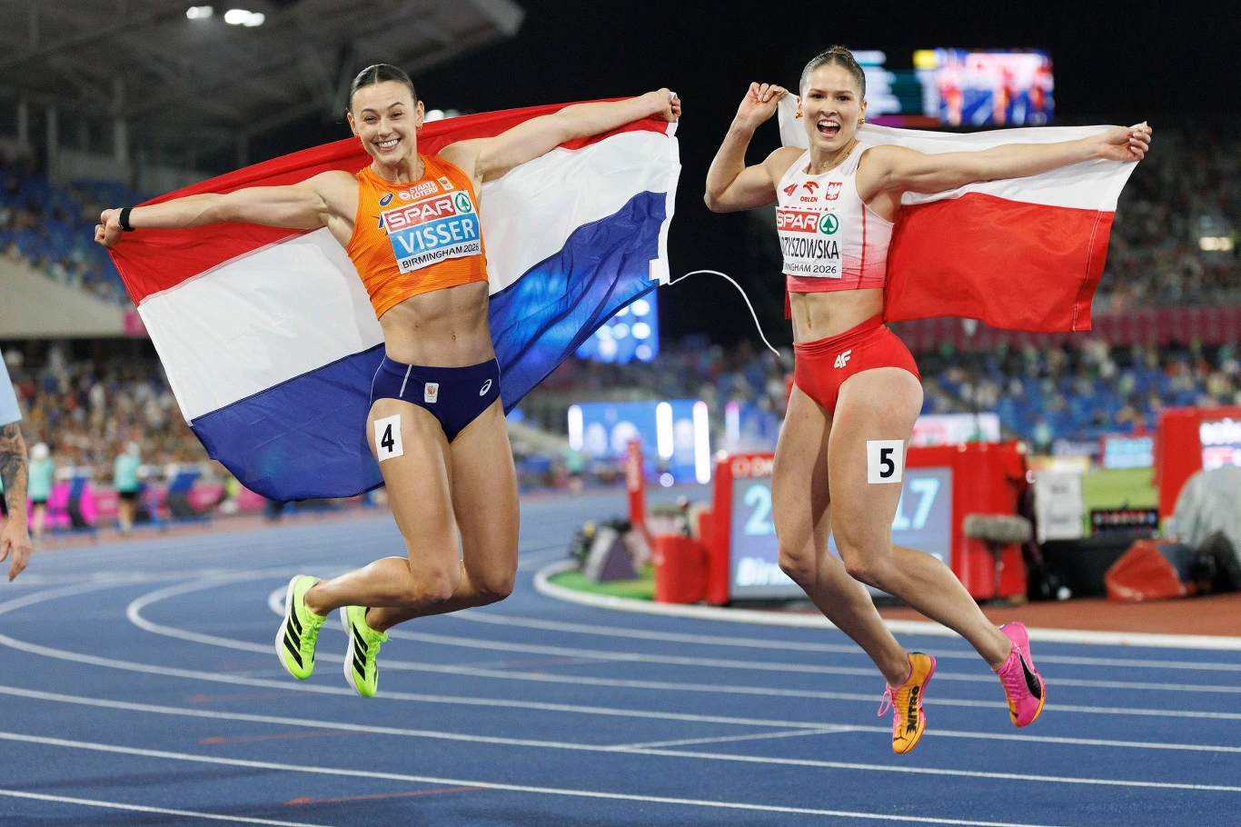 Pia Skrzyszowska i Nadine Visser Dwie lekkoatletki w strojach reprezentacyjnych w trakcie radosnego skoku na bieżni z rozpostartymi flagami swoich krajów, z wyraźnymi numerami startowymi na udach.