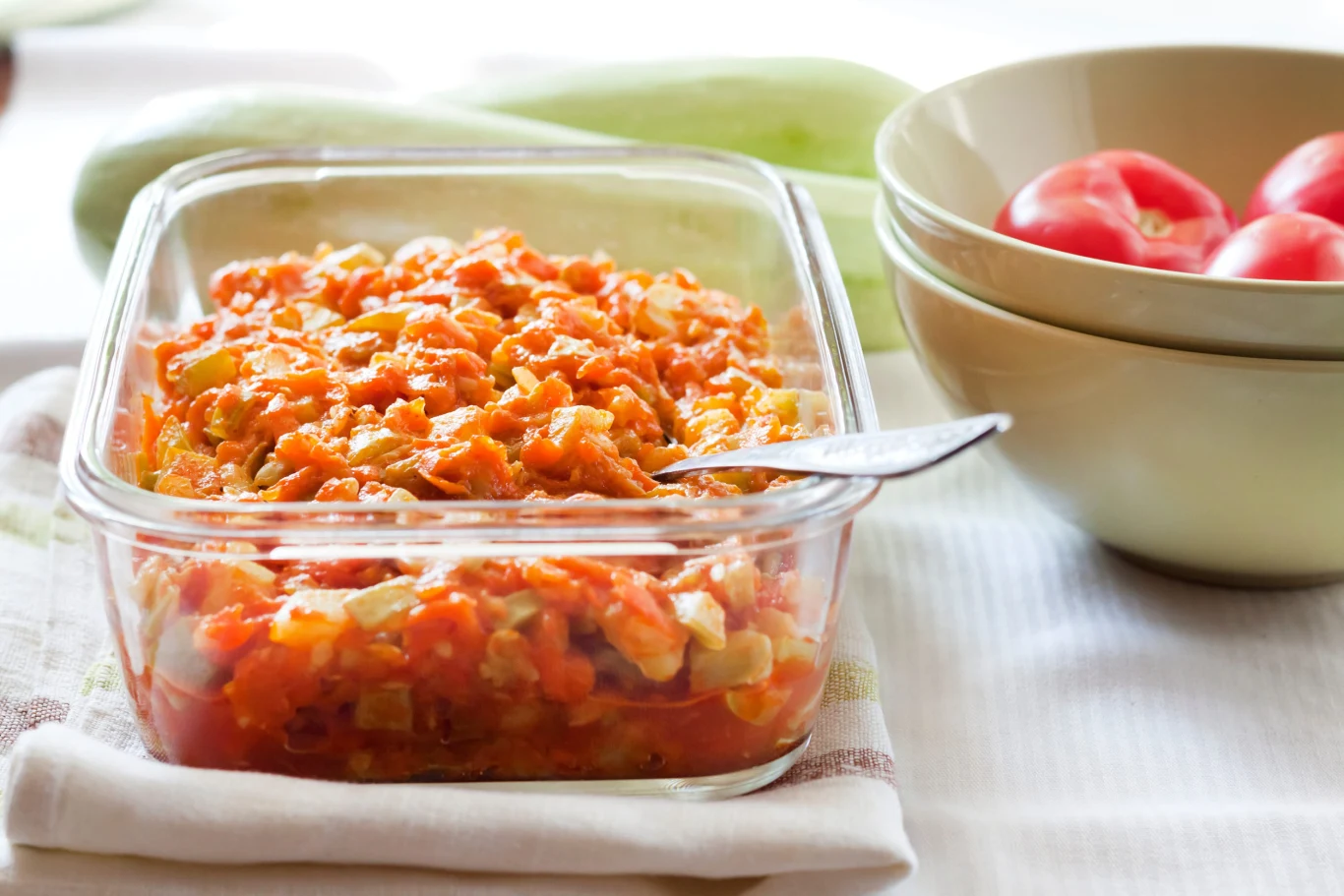 Bigos z cukinii zaskakuje smakiem. Warto wypróbować ten przepis Szklane naczynie z bigosem z cukinii w sosie pomidorowym, obok miska z czerwonymi pomidorami na ściereczce.