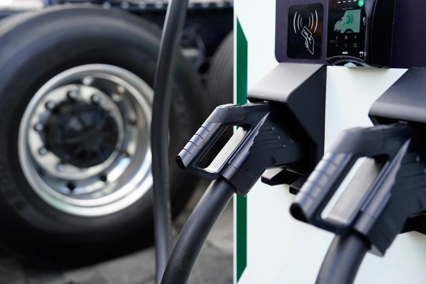 Rozwój rynku elektrycznych ciężarówek przebiega wolniej niż oczekiwano Dwie końcówki ładowarki do pojazdów elektrycznych zamocowane w stacji ładowania na tle koła ciężarówki.