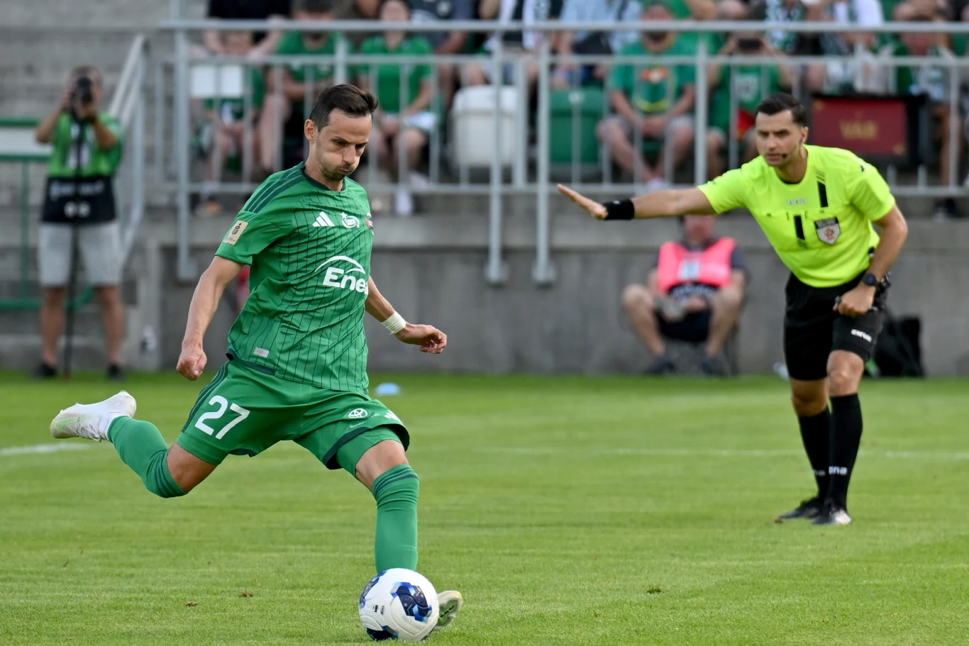 Rafał Wolski w barwach Radomiaka Radom Piłkarz w zielonym stroju przygotowujący się do kopnięcia piłki podczas meczu, obok niego sędzia wykonujący gest wskazujący kierunek gry.