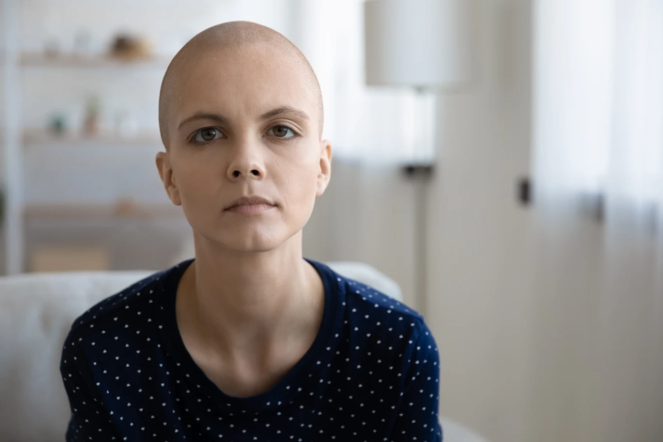 Chemo brain może powodować problemy z pamięcią, koncentracją i szybkością myślenia podczas leczenia onkologicznego lub po jego zakończeniu Młoda osoba z ogoloną głową w granatowej bluzce w białe kropki siedzi na sofie, w tle jasny pokój.