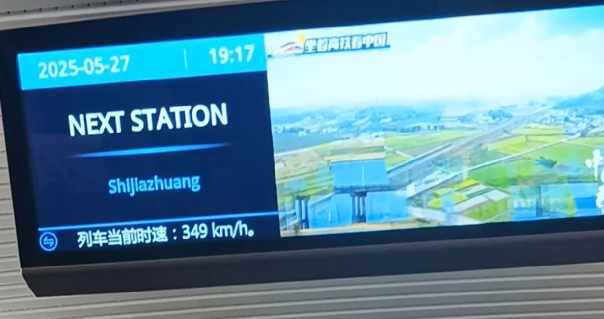 Zapowiedź nadjeżdżającego pociągu Ekran informacyjny w pociągu pokazujący aktualną prędkość 349 km/h, godzinę 19:17, datę 27 maja 2025 roku oraz informację o następnej stacji Shijiazhuang, obok zdjęcie krajobrazu z torami kolejowymi i polami.