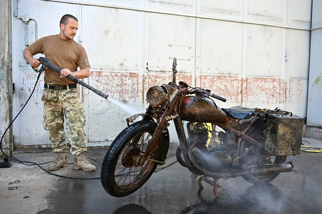 Odnaleziony DKW zostanie odrestaurowany. mężczyzna w wojskowych spodniach używa myjki ciśnieniowej do czyszczenia zabłoconego i zardzewiałego motocykla ustawionego na betonowej powierzchni