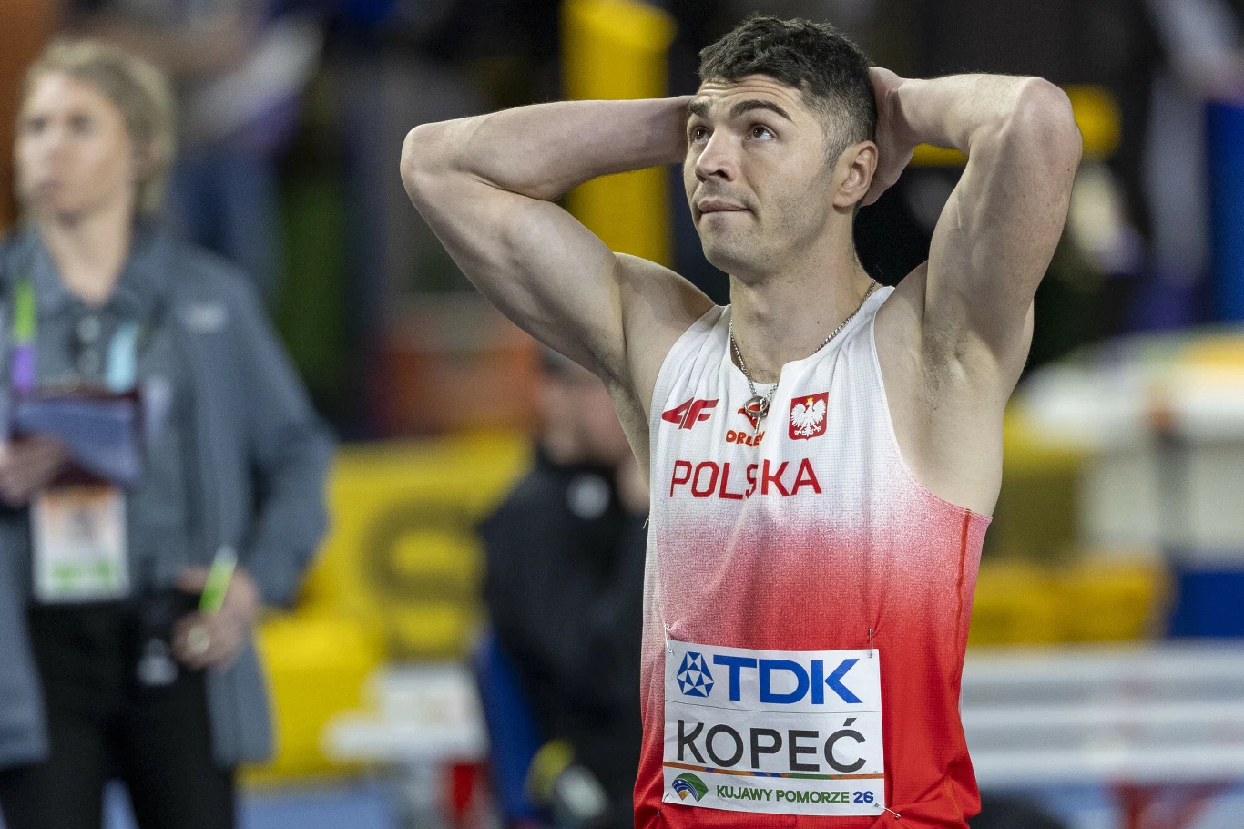 Lekkoatleta w koszulce reprezentacji Polski z numerem startowym Kopeć, stojący z rękami za głową i wyrazem skupienia na twarzy na tle hali sportowej.