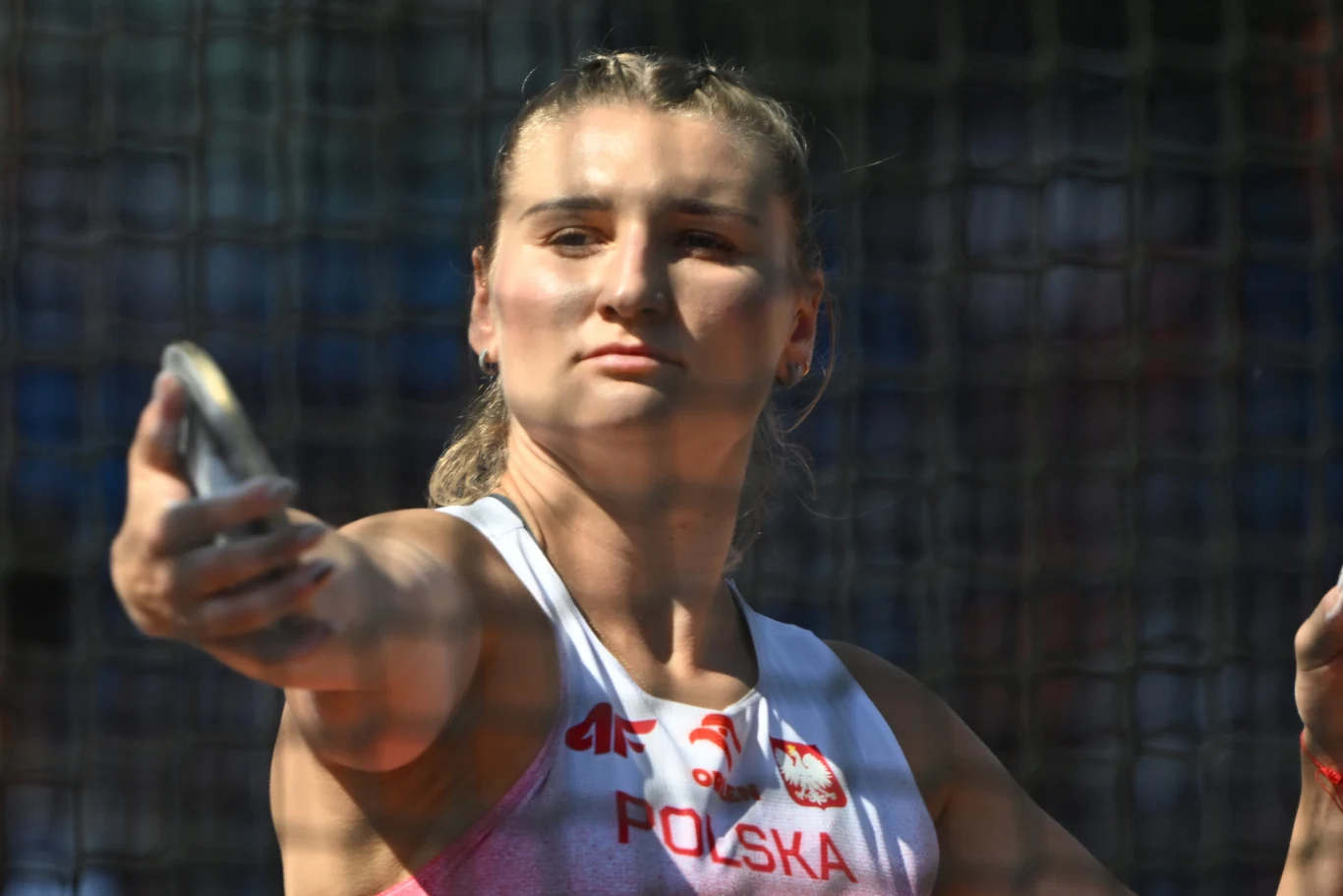 Daria Zabawska Lekkoatletka w polskim stroju sportowym skupiona przed rzutem dyskiem, widoczna zza siatki ochronnej stadionu.