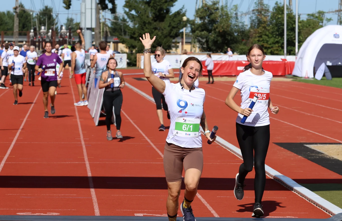 Grupa uczestników biegu sztafetowego na stadionie lekkoatletycznym, uśmiechnięta kobieta z pałeczką unosi rękę w geście radości, obok młoda zawodniczka biegnąca w stronę mety.
