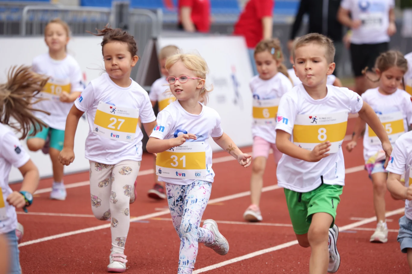 Grupa kilku uśmiechniętych dzieci w białych koszulkach i kolorowych numerach startowych biegnie po bieżni stadionu sportowego.