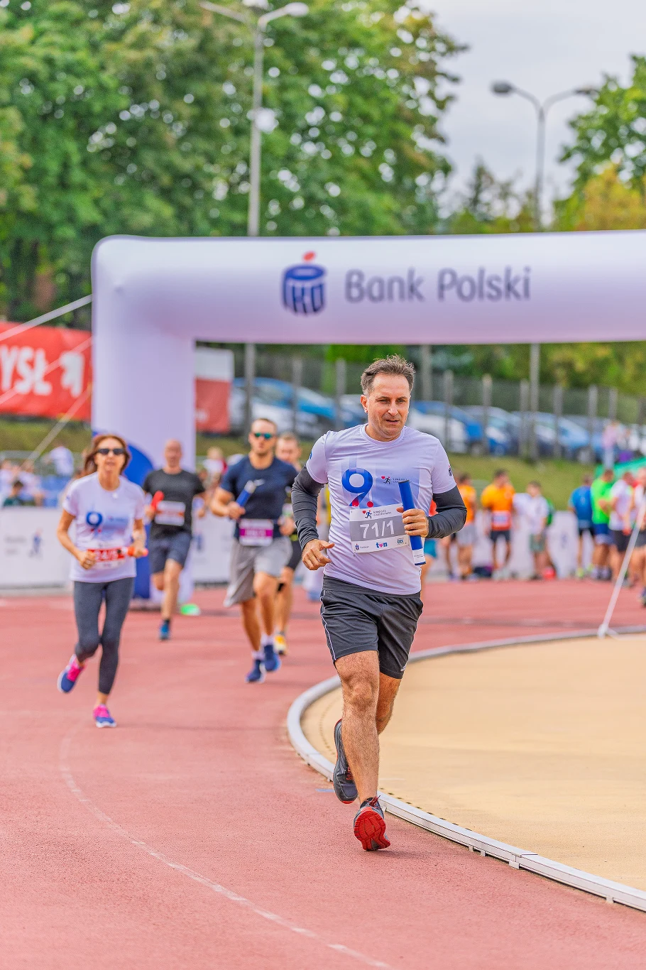 Grupa biegaczy ubranych w sportową odzież pokonuje dystans na stadionie lekkoatletycznym, przechodząc pod dużym, białym łukiem z logo PKO Banku Polskiego.