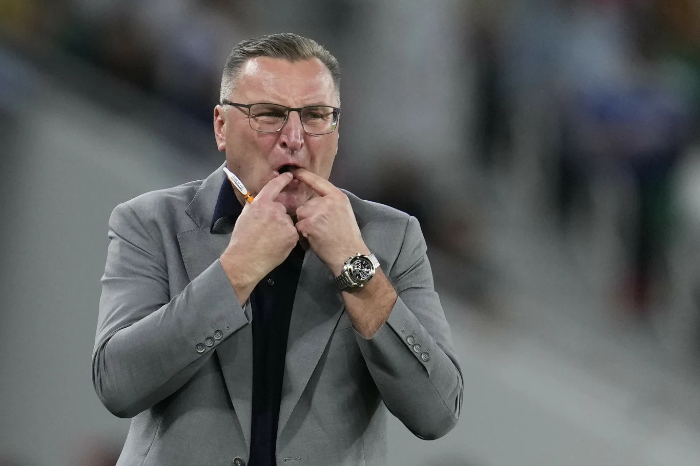 Czesław Michniewicz Czesław Michniewicz w szarej marynarce i okularach gwiżdże na palcach na tle trybun stadionu.