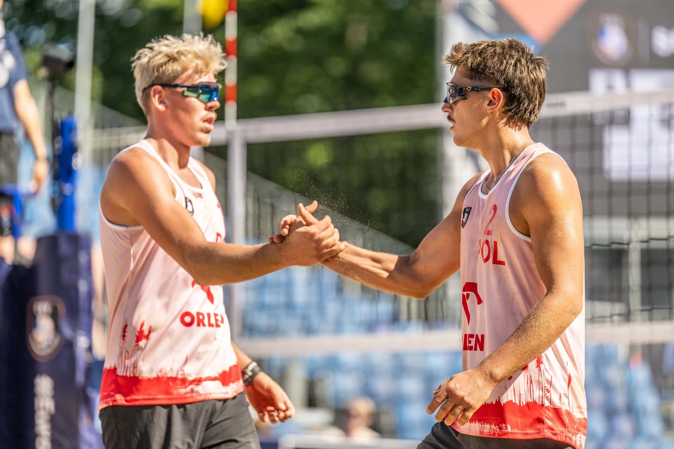 Szymon Beta i Artem Besarab Dwóch młodych siatkarzy plażowych w sportowych strojach ściska sobie dłonie podczas meczu, obaj mają na sobie okulary przeciwsłoneczne i koszulki z logo sponsoringowym, w tle widoczna siatka oraz fragment trybun.