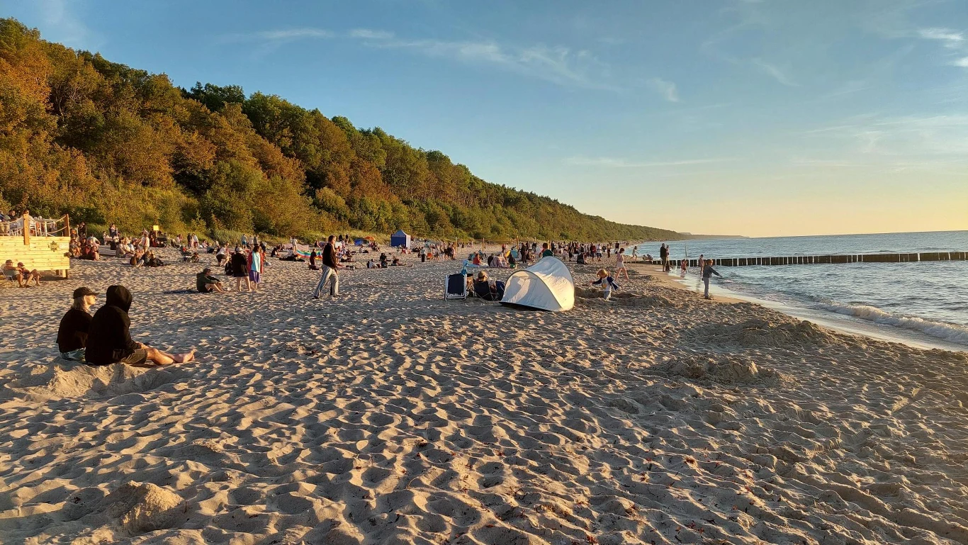 Plaża o zachodzie słońca z wieloma osobami odpoczywającymi na piasku, białym namiotem i wałem zielonych drzew w tle, spokojne morze po prawej stronie kadru.