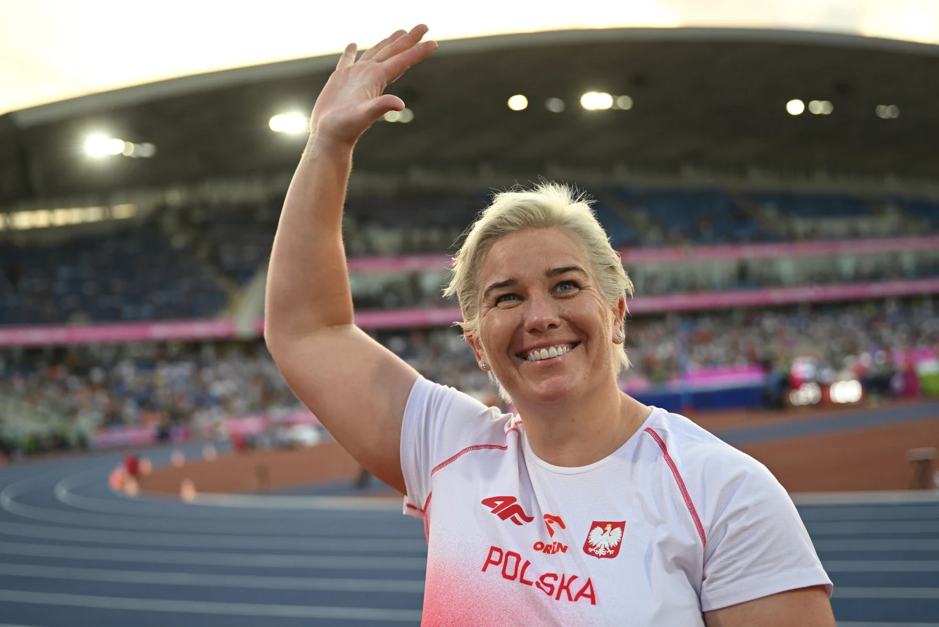 Anita Włodarczyk Uśmiechnięta kobieta w białej koszulce z napisem 'POLSKA' i godłem, podnosząca rękę w geście powitania na stadionie lekkoatletycznym.